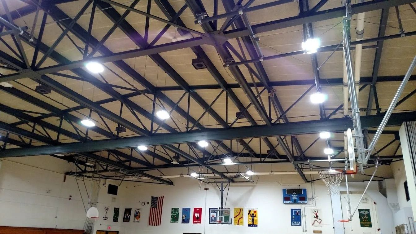 Basketball court interior, featuring a truss ceiling with lights and a basketball hoop. Banners hang on the wall.