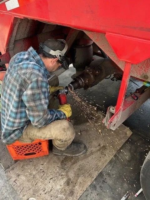 Mechanic in plaid shirt uses a power tool under a red vehicle, with metal shavings on the floor.