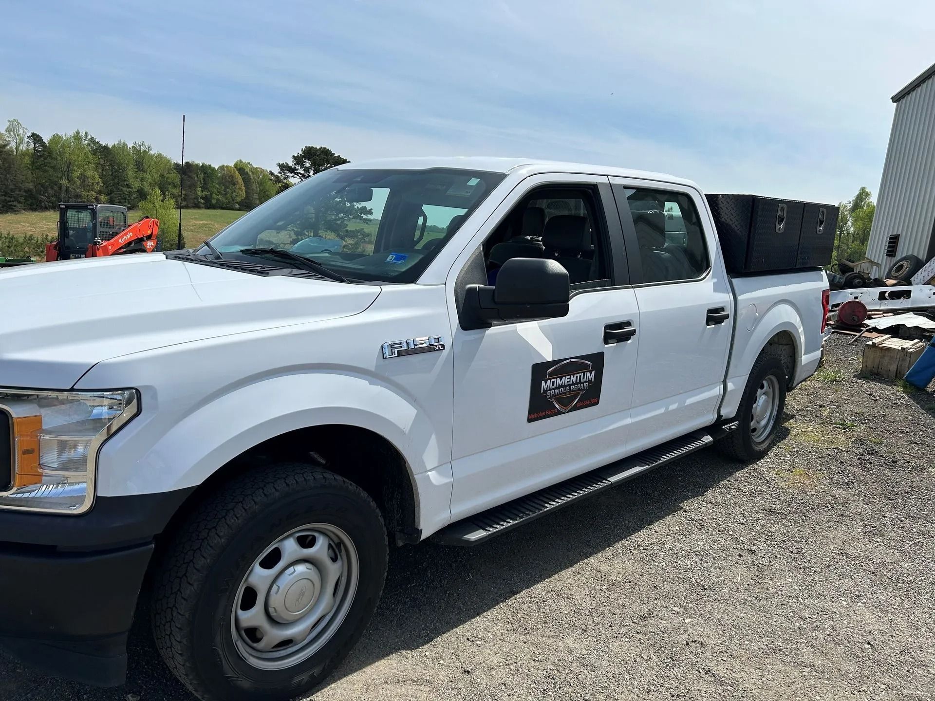 White pickup truck parked on gravel beside an industrial yard, with a black sign on the door and open rear bed.