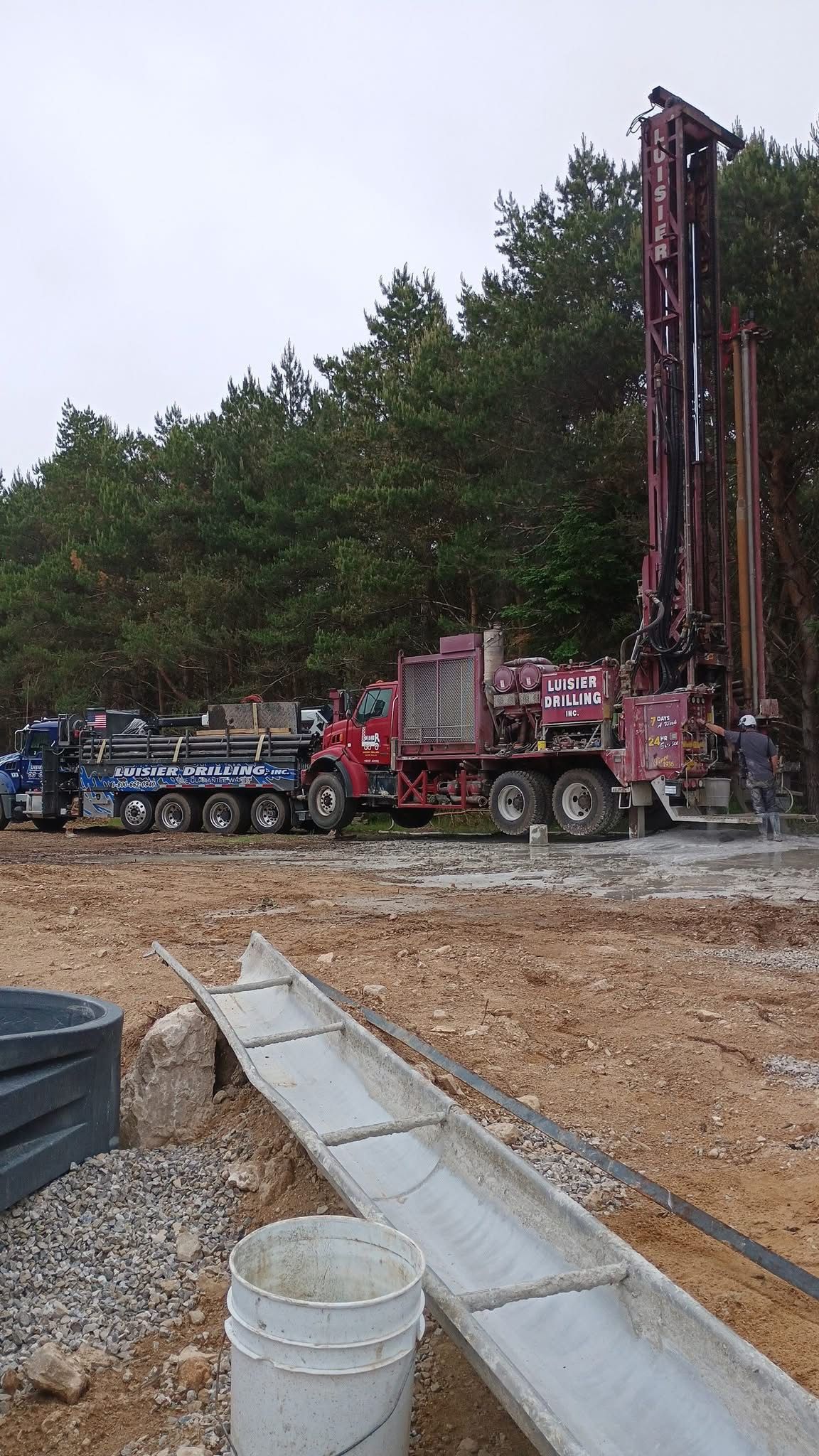 Drilling rig and support trucks on a muddy construction site beside trees and a gravel trench