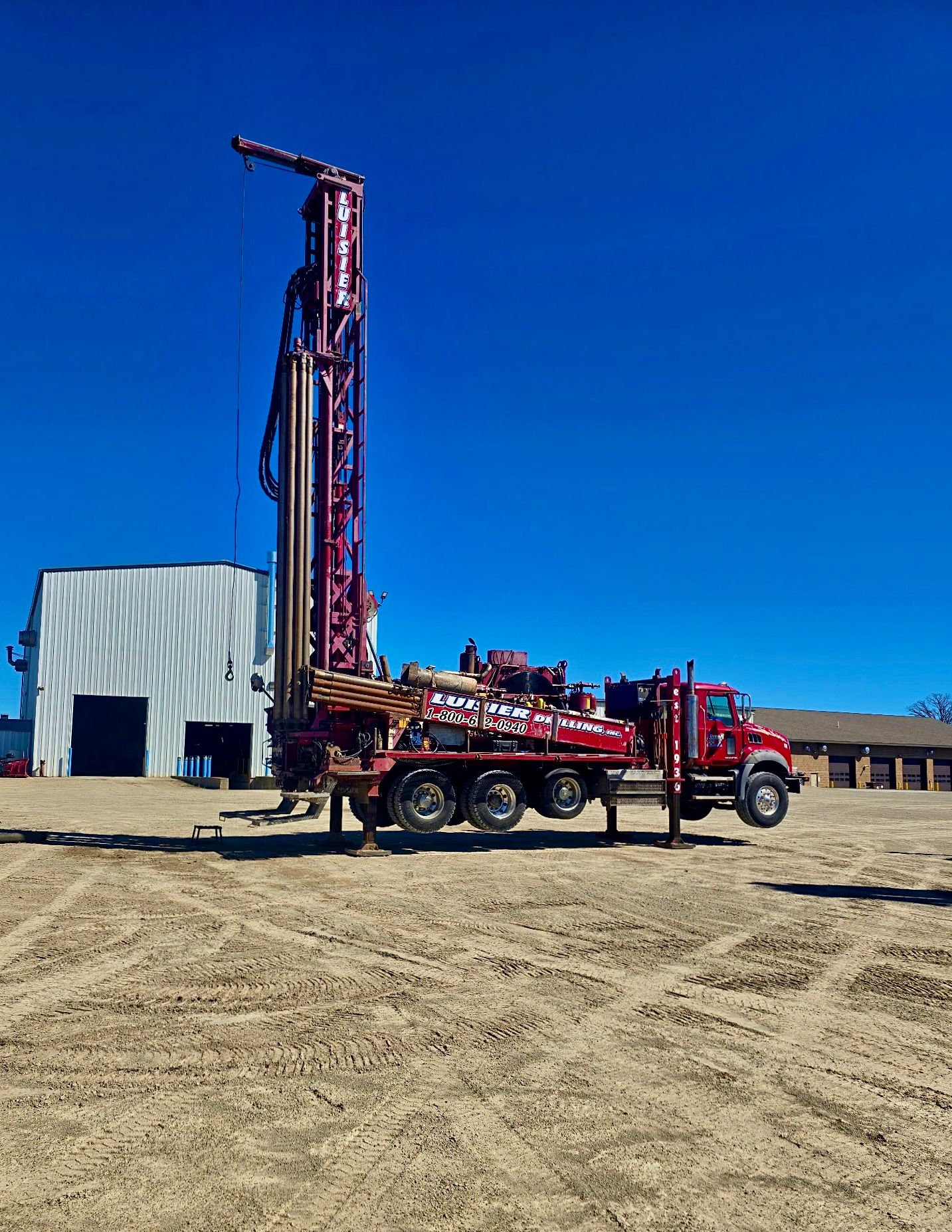 Red drilling rig truck parked on a dirt lot beside a small metal building under a clear blue sky