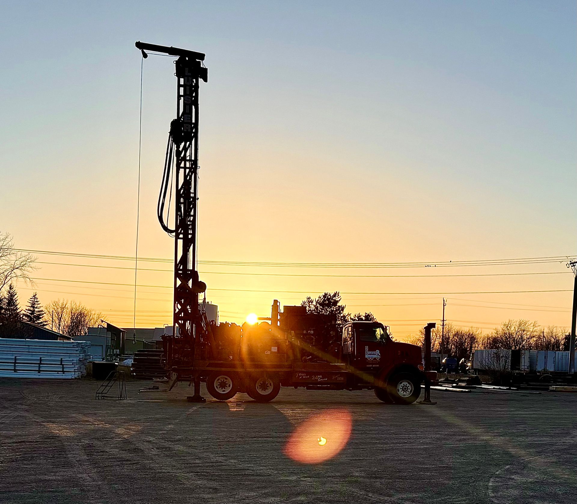 Drilling rig silhouetted at sunset on a gravel lot, with orange glow and a clear horizon.