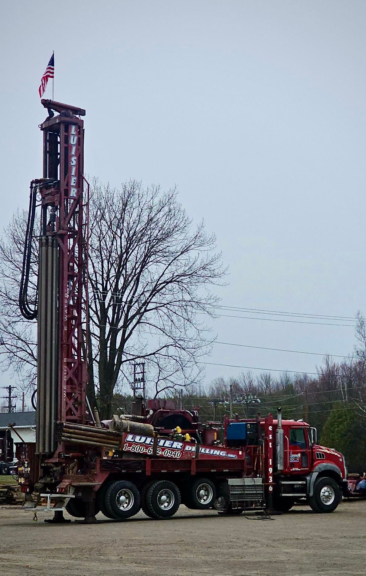 Red drilling rig truck with extended mast parked outdoors under a gray sky