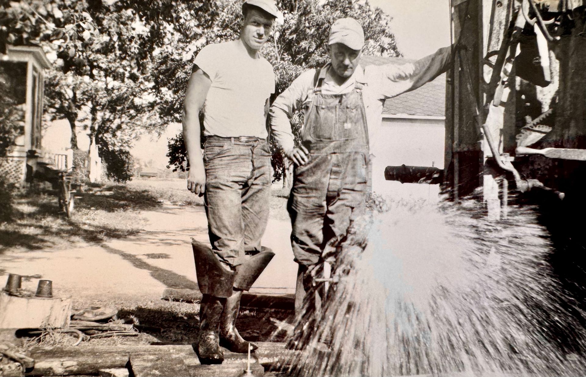 Two workers in boots and hats stand by a spraying hose or water jet outdoors near a building.