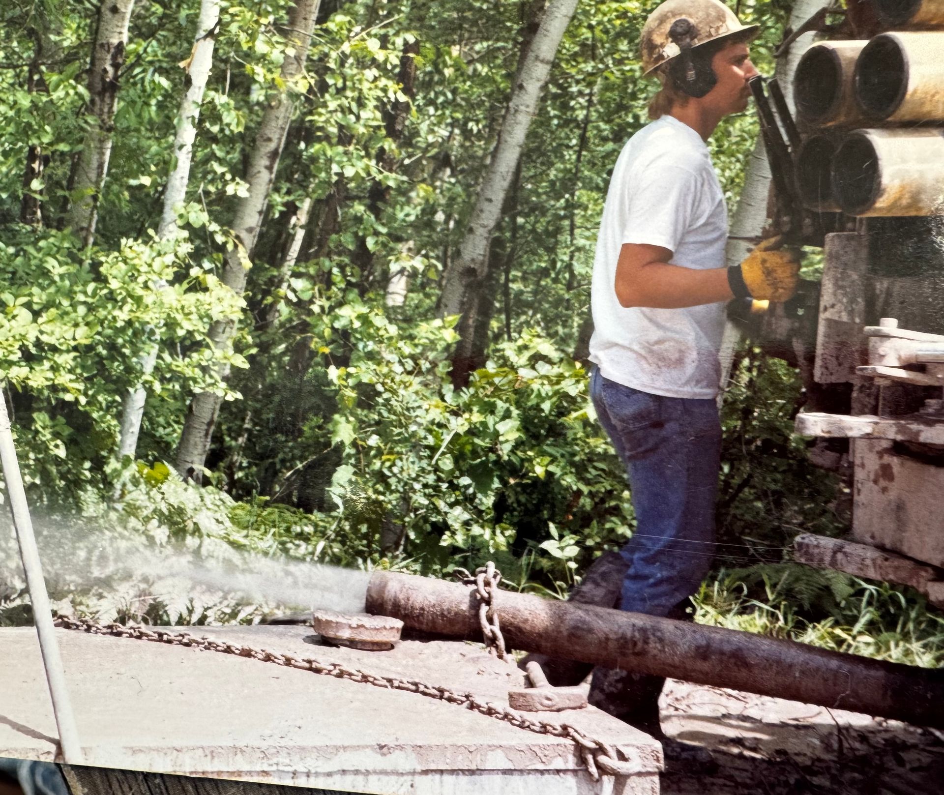 Worker in hard hat and gloves standing by stacked pipes outdoors near a hose spraying water
