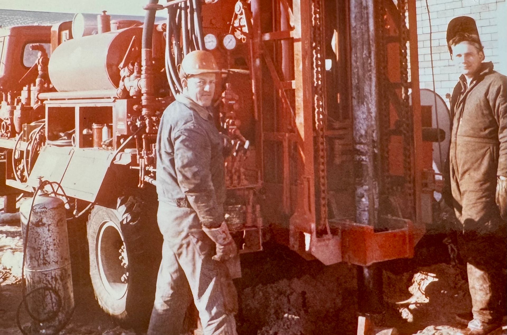 Two workers stand beside a red industrial vehicle or machine in a dusty outdoor worksite.