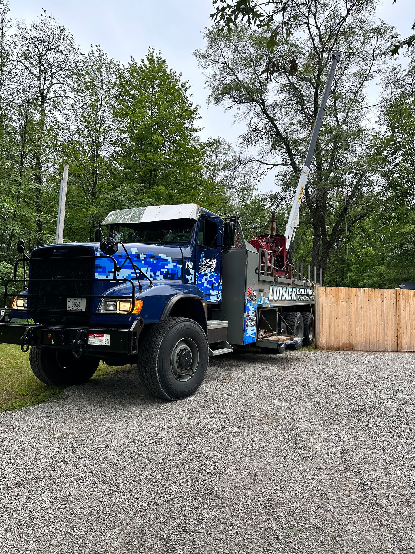 Blue tow truck with a person standing in the back, parked on gravel beside a wooden fence and trees.