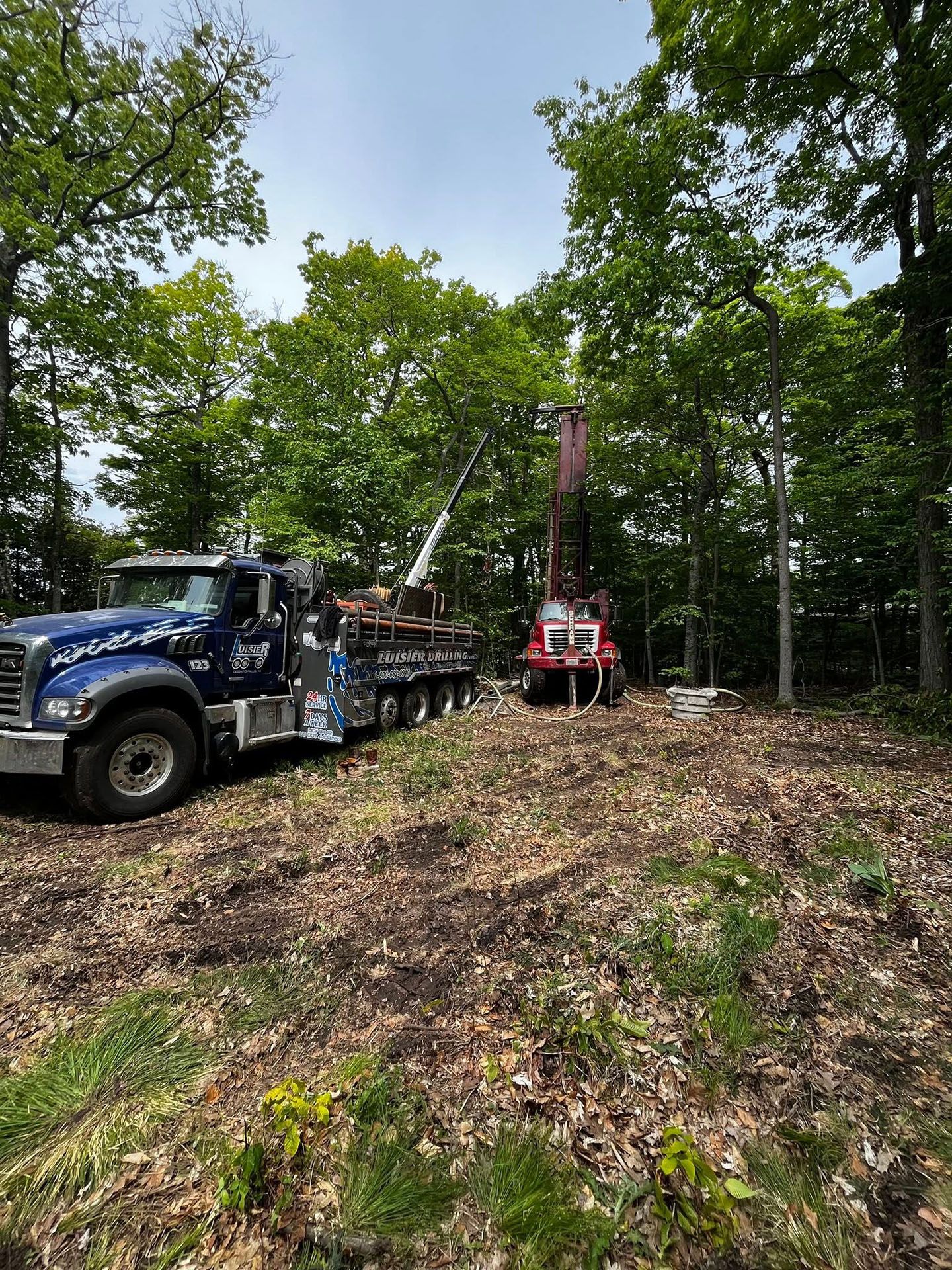Blue truck and red drilling rig in a wooded clearing with leaf-covered ground