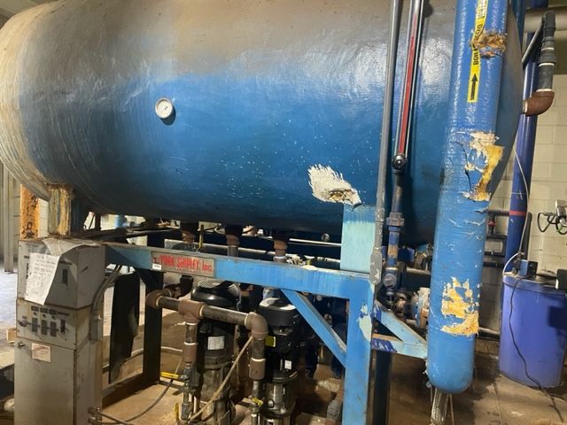 Blue industrial tank with pipes and gauges on a metal frame, in an industrial setting.