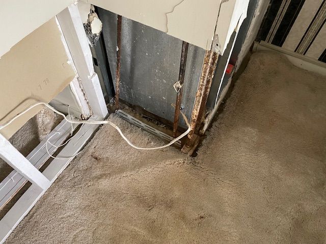 Damaged wall with exposed pipes and carpet. Rusted metal, crumbling plaster, and loose electrical wire.