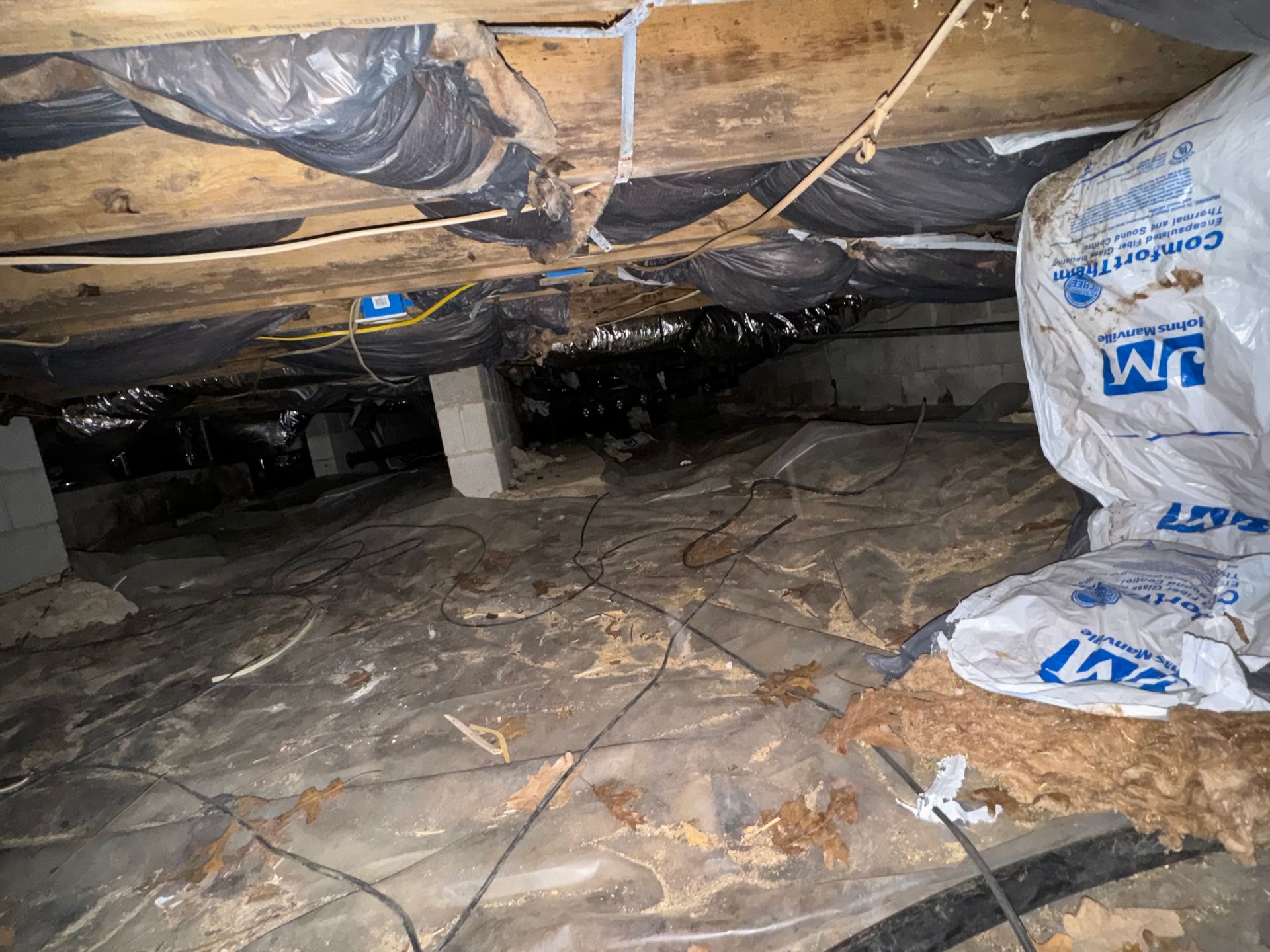 Crawl space beneath a house with insulation, plastic sheeting on the ground, and electrical wiring.