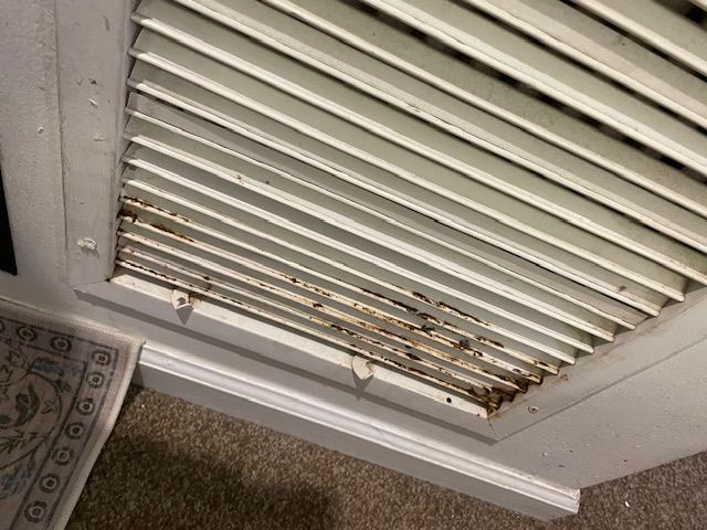 White air vent with rust and debris, set in a white frame, attached to a wall.