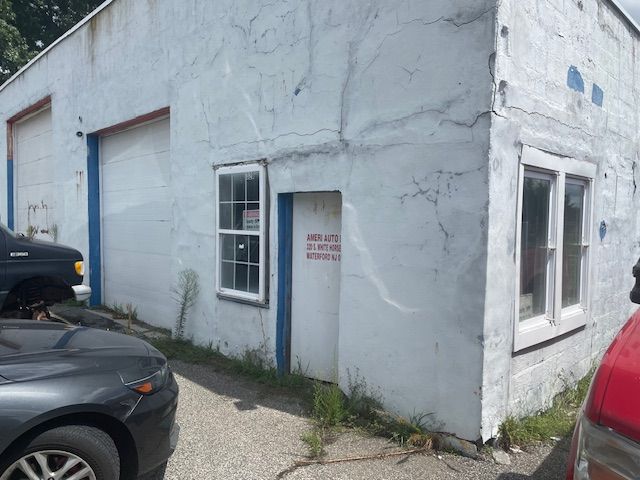 Exterior of a rundown auto shop. White building with roll-up doors, windows, and cracked stucco. Cars parked nearby.