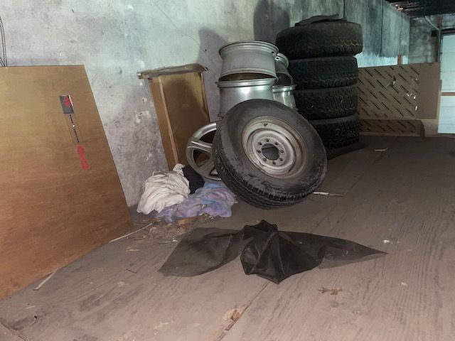 Tires stacked in a cluttered basement corner with plywood, a trash can, and auto parts.