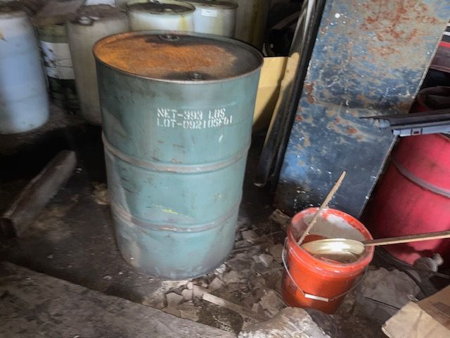 Green metal drum, partially rusted, indoors. A bucket with a wooden spoon sits nearby.