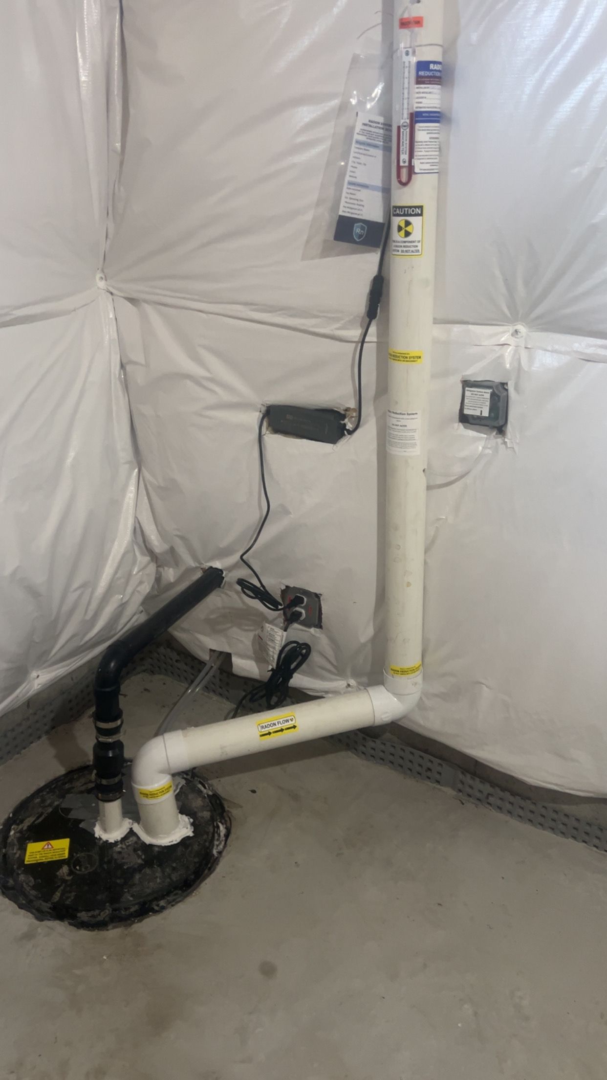 Sump pump system in a basement with white insulation. Black and white pipes, a pump, and electrical components are visible.