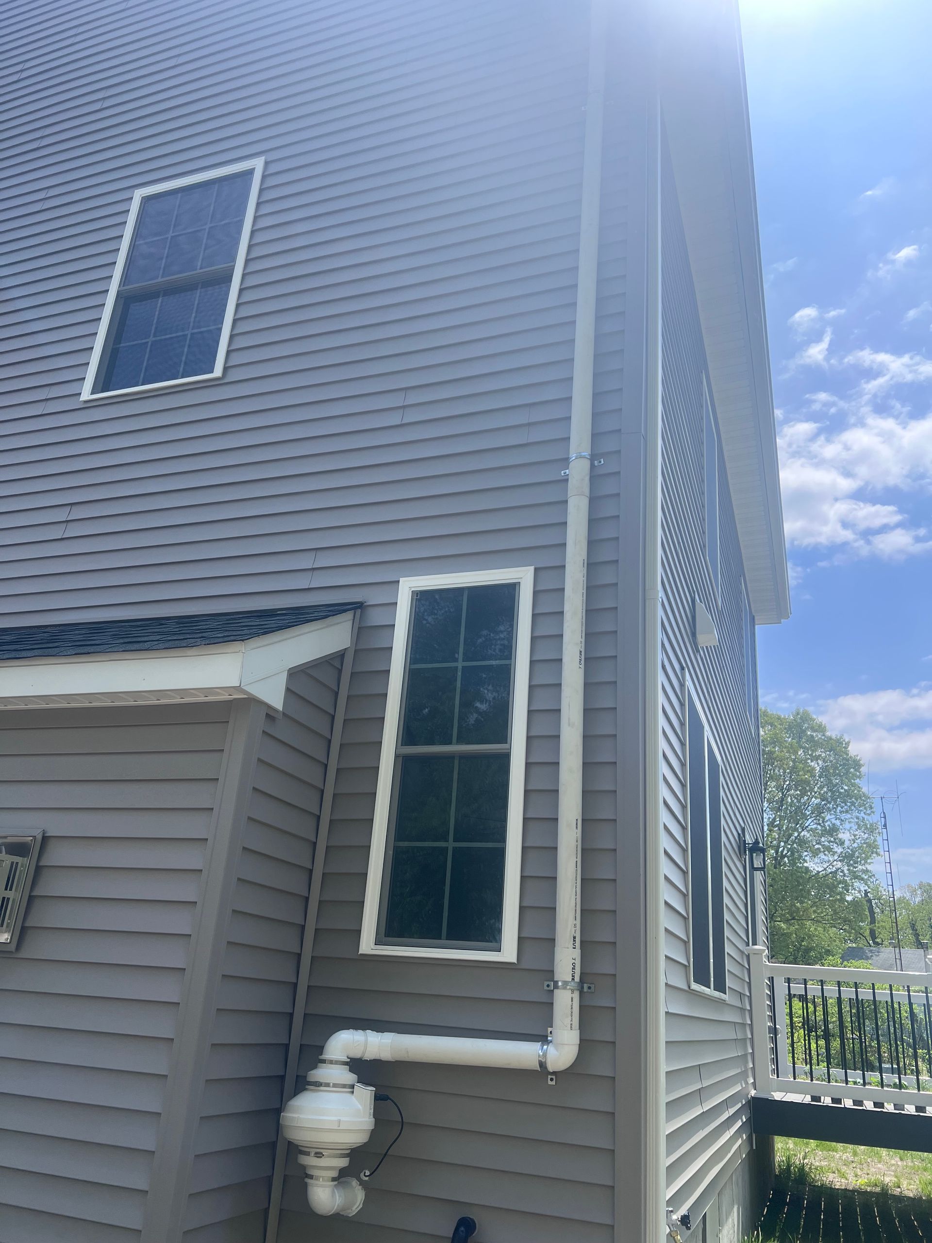 Side of a house with gray siding, windows, and white pipes against a blue sky.