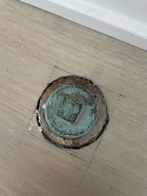 A circular, teal-colored metal access cover set into a wooden floor with a square-shaped opening, surrounded by worn wood and flooring.