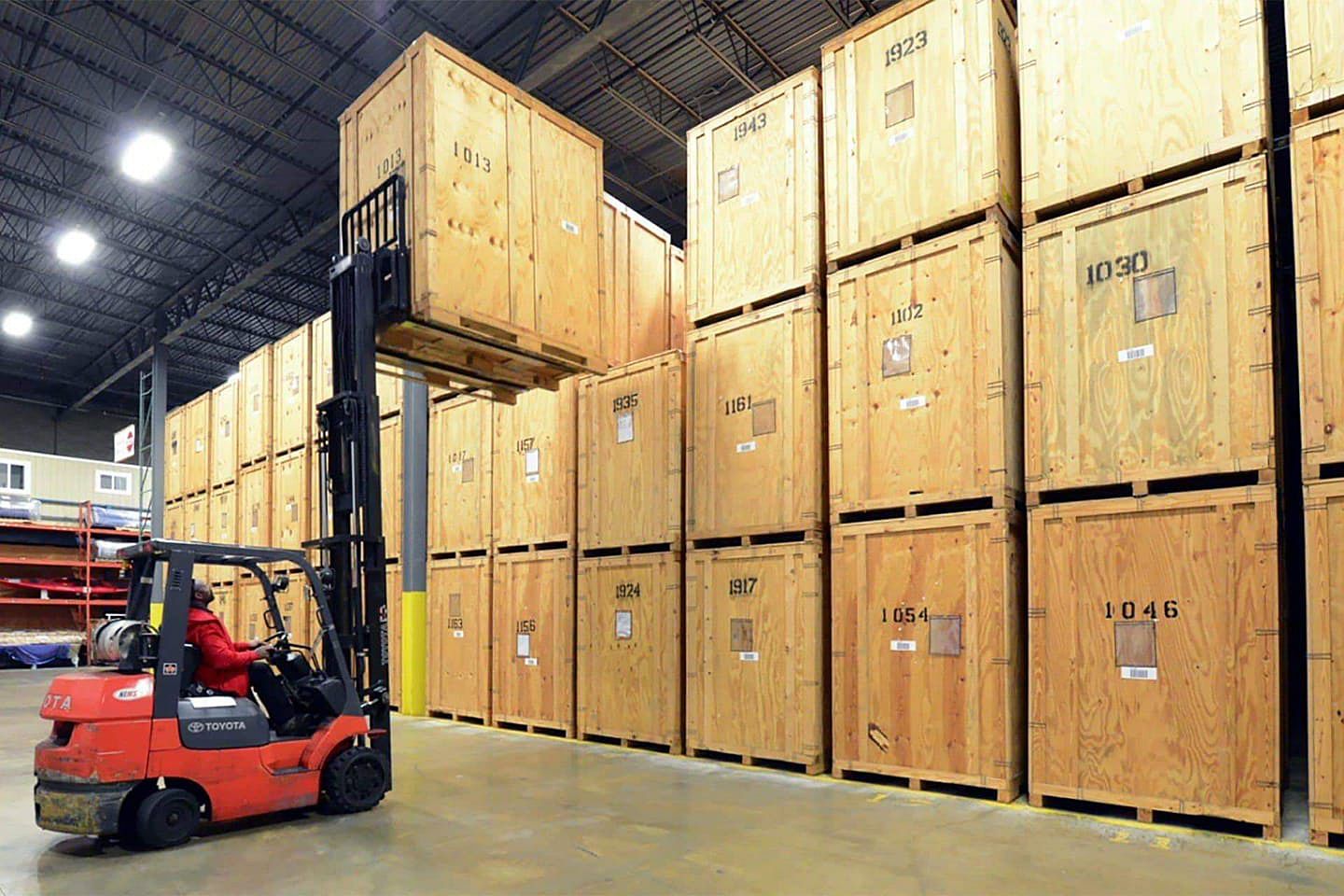 Forklift lifting a wooden crate in a storage warehouse; many stacked wooden crates line the wall.