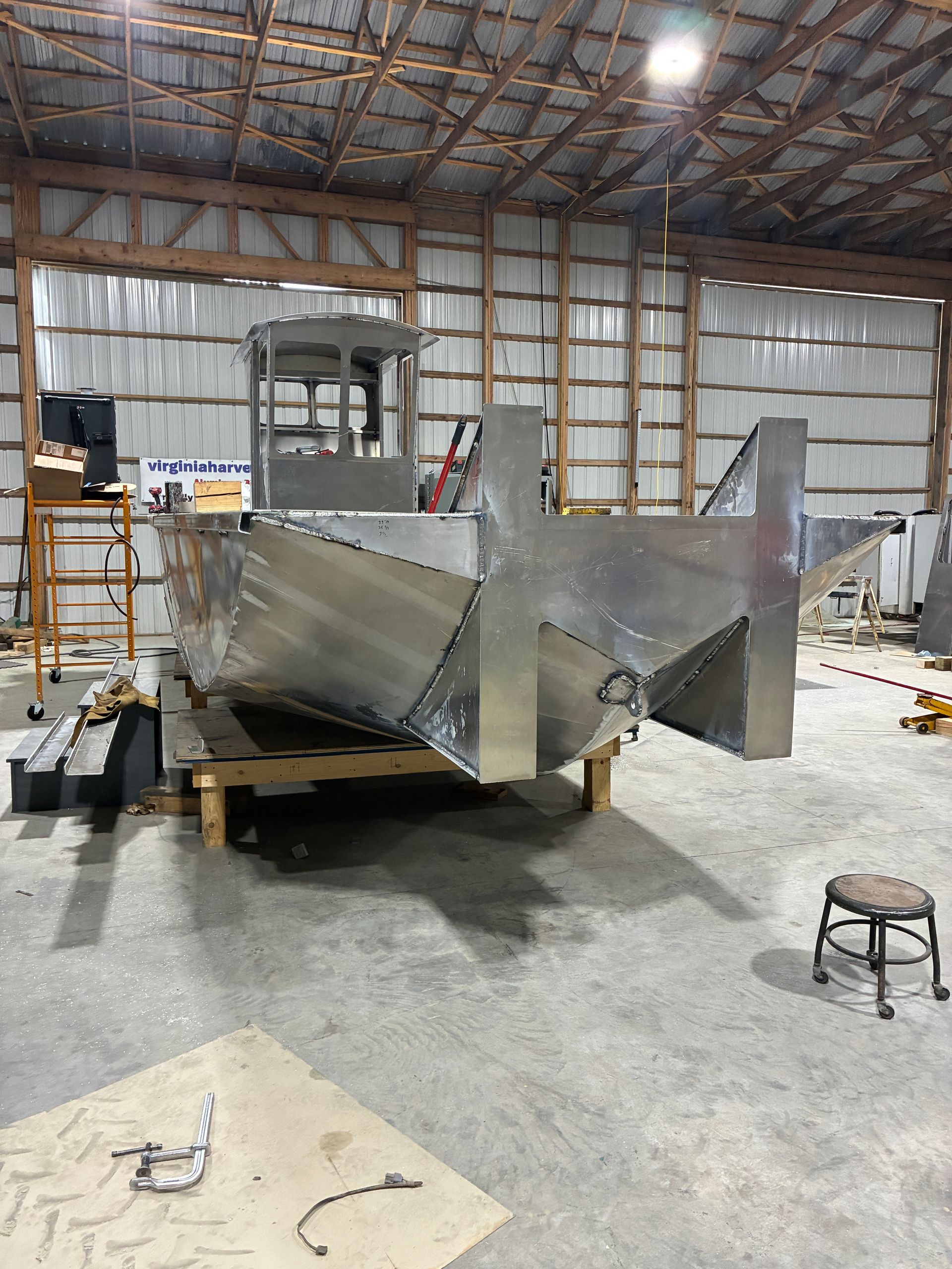An aluminum boat hull under construction, elevated on wooden blocks inside a large, industrial metal workshop.