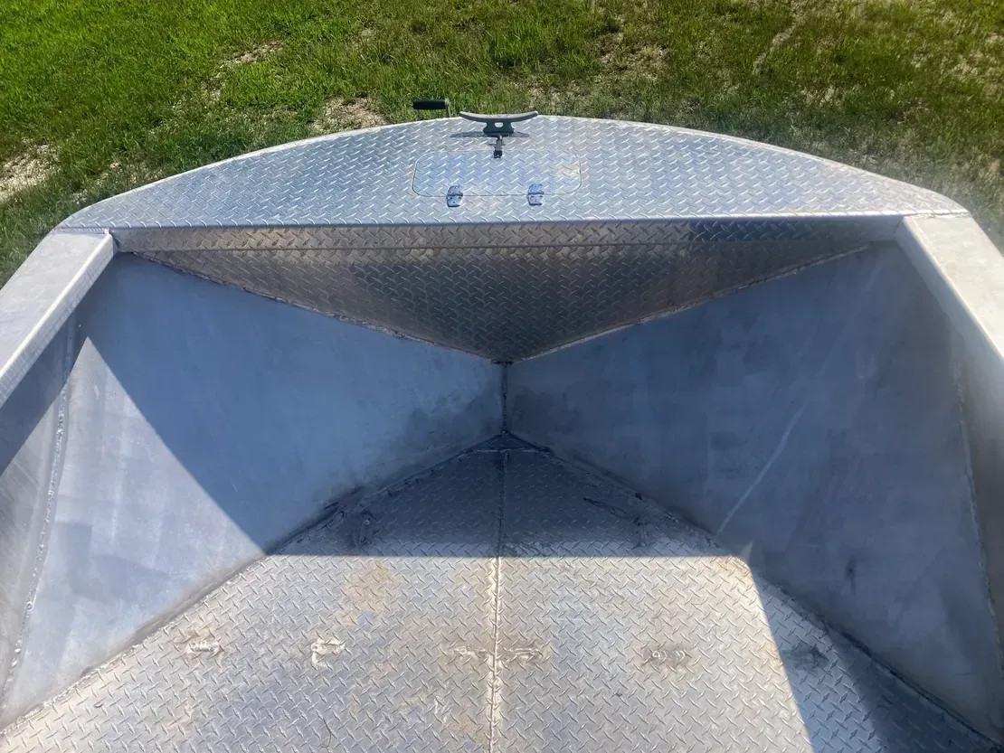 The bow of a metal boat showing a diamond-plate deck, storage compartment, and mooring cleat in a grassy outdoor setting.