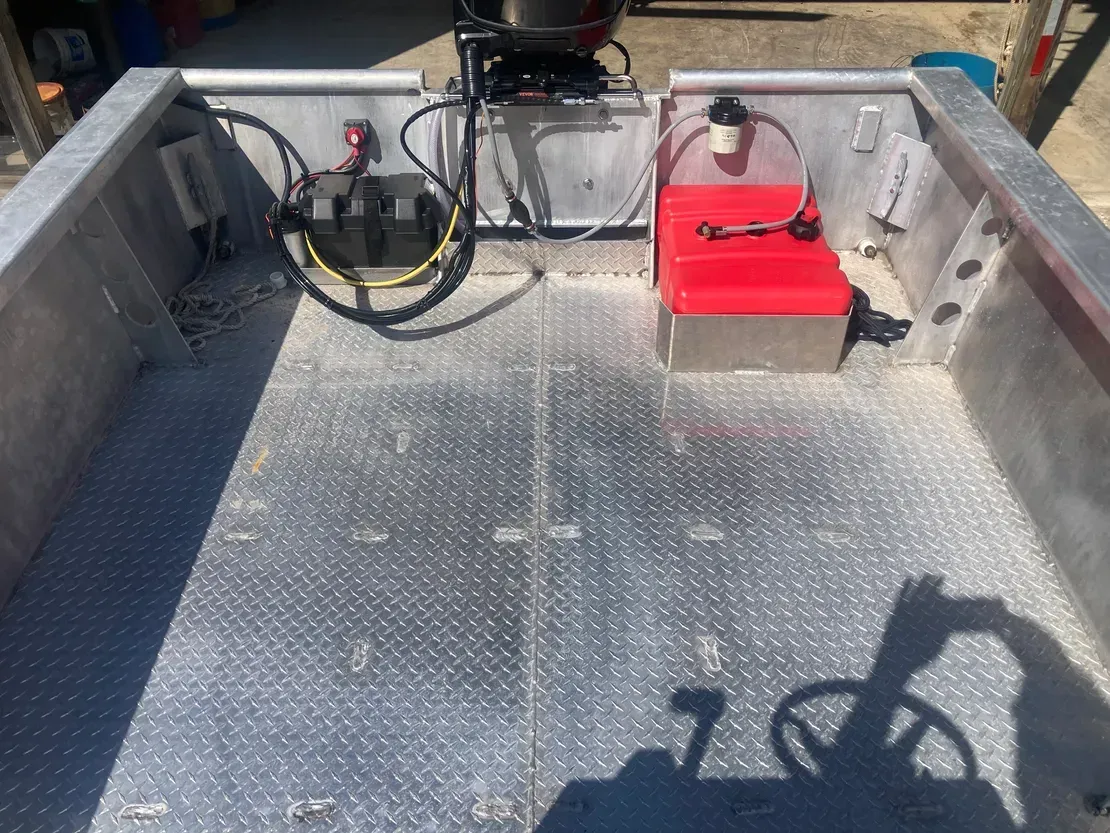Rear interior of a metal boat featuring a diamond-plate floor, a black battery, and a red fuel tank with fuel lines.
