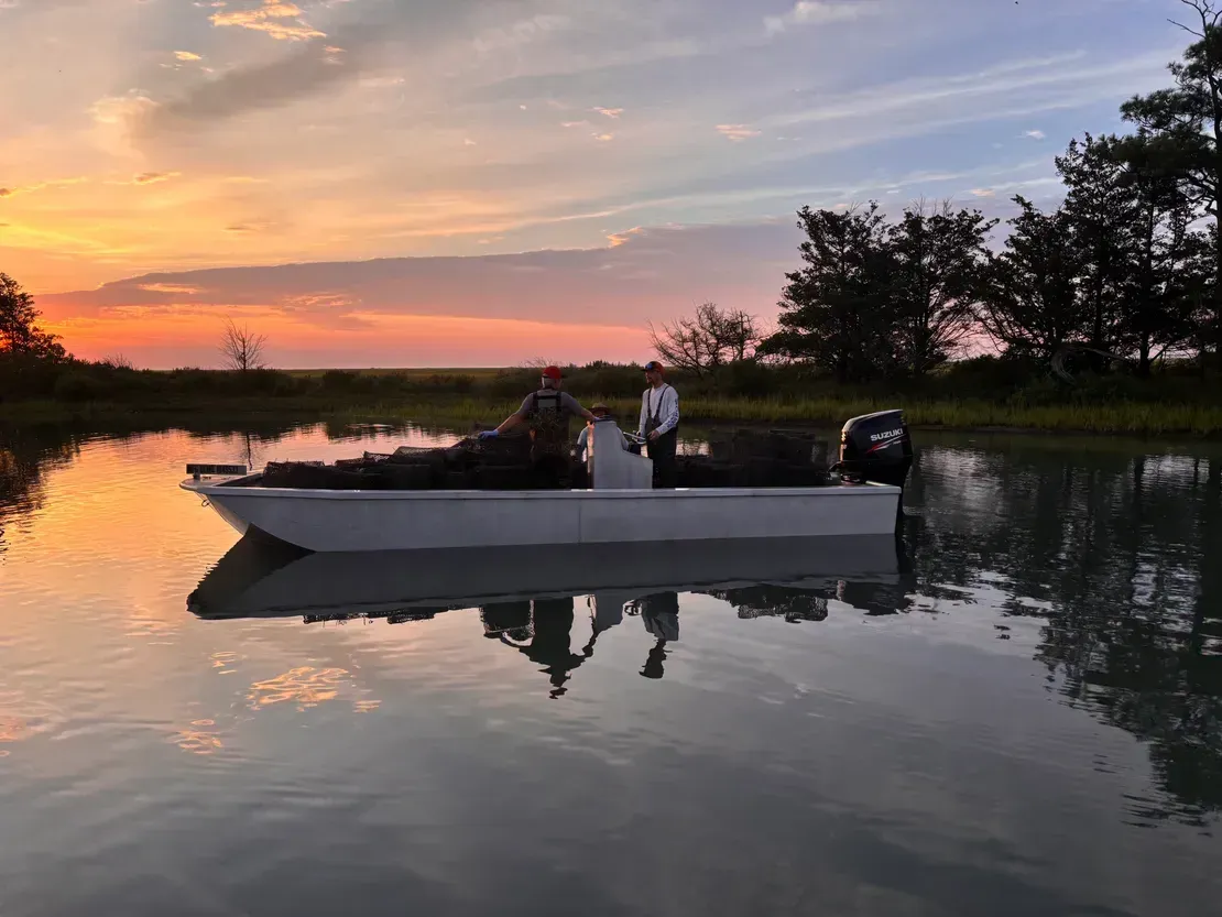 Two people in a small boat on calm, reflective water at sunset, with coastal marshland in the background.