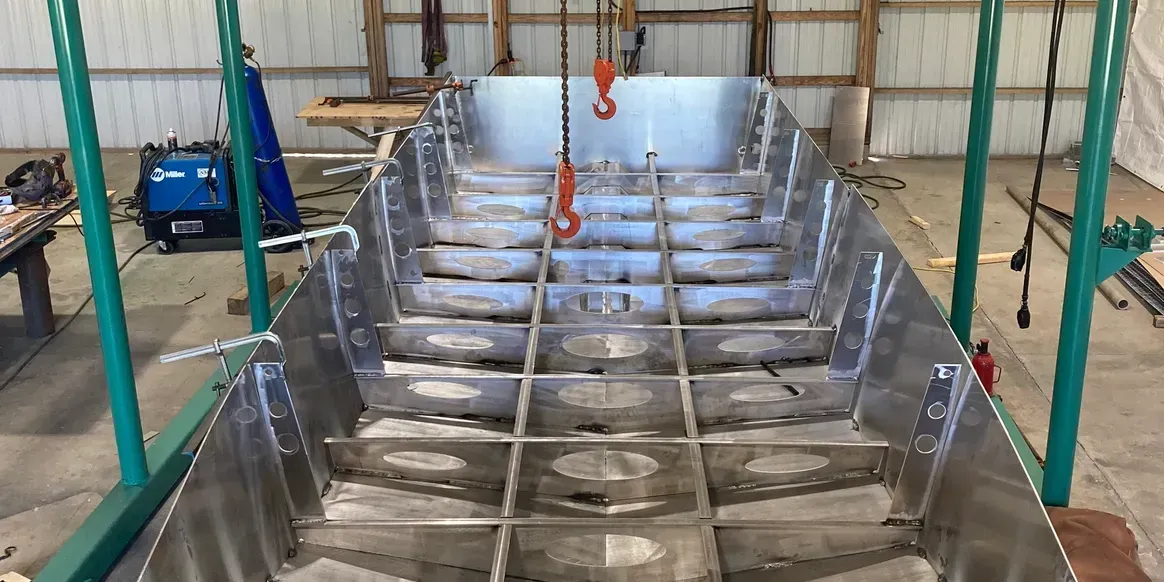 The interior structural frame of a metal boat hull under construction in a workshop, featuring a grid of welded ribs.