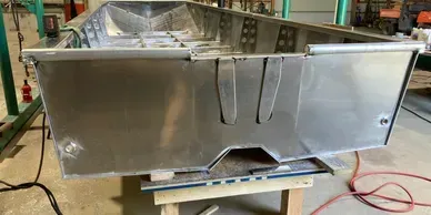 An unfinished aluminum boat hull sits on a wooden workbench inside a metalworking shop.