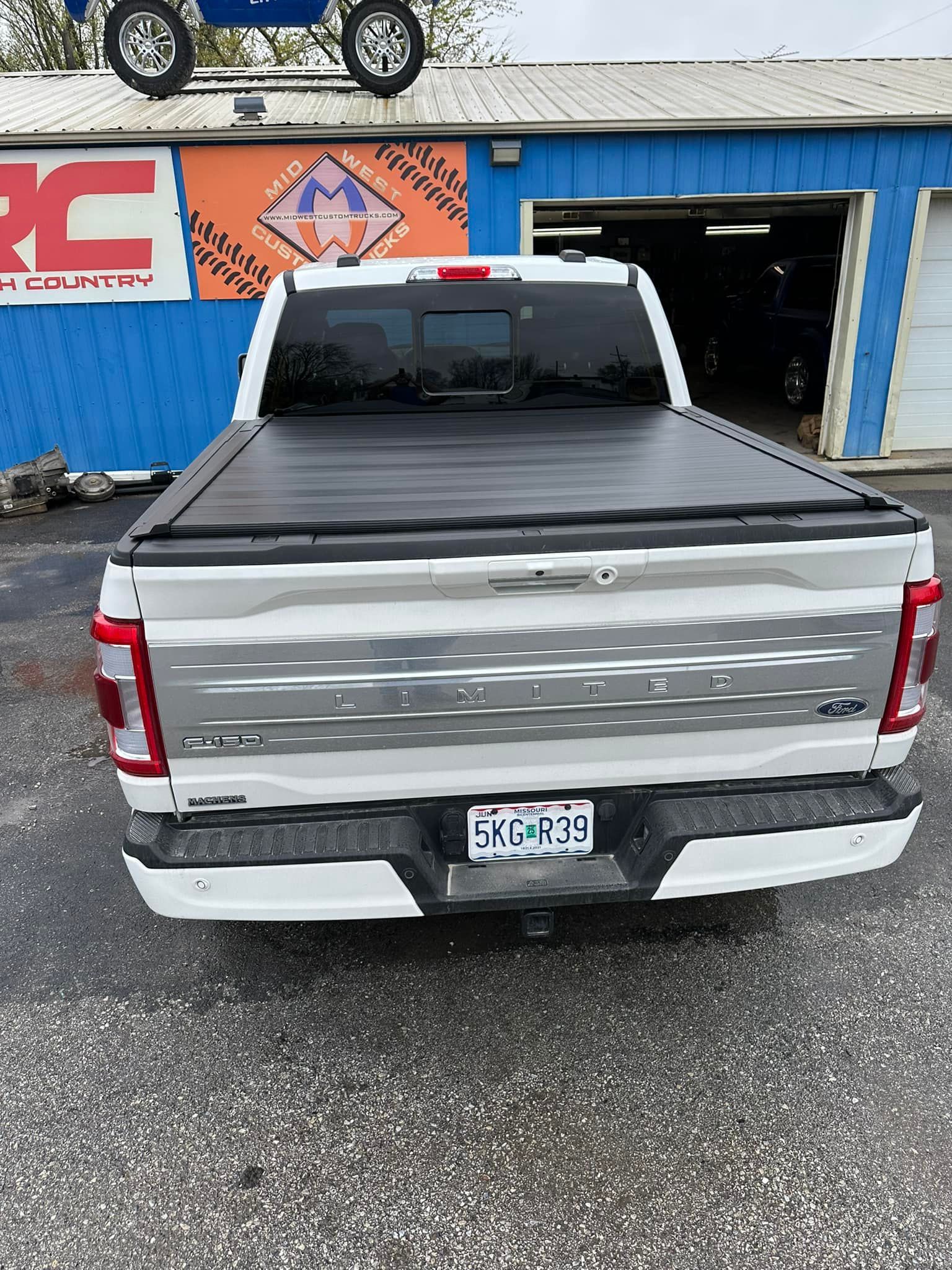 A white truck with a black tonneau cover is parked in front of a blue building.