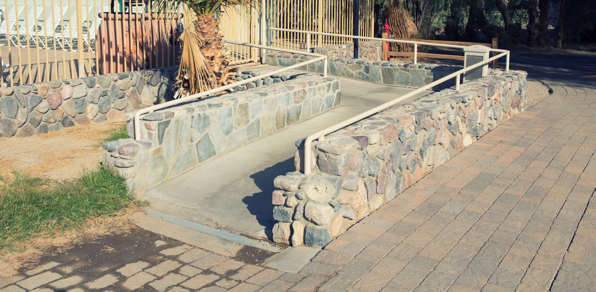 Stone ramp with handrails leading to a building, palm tree in the background.