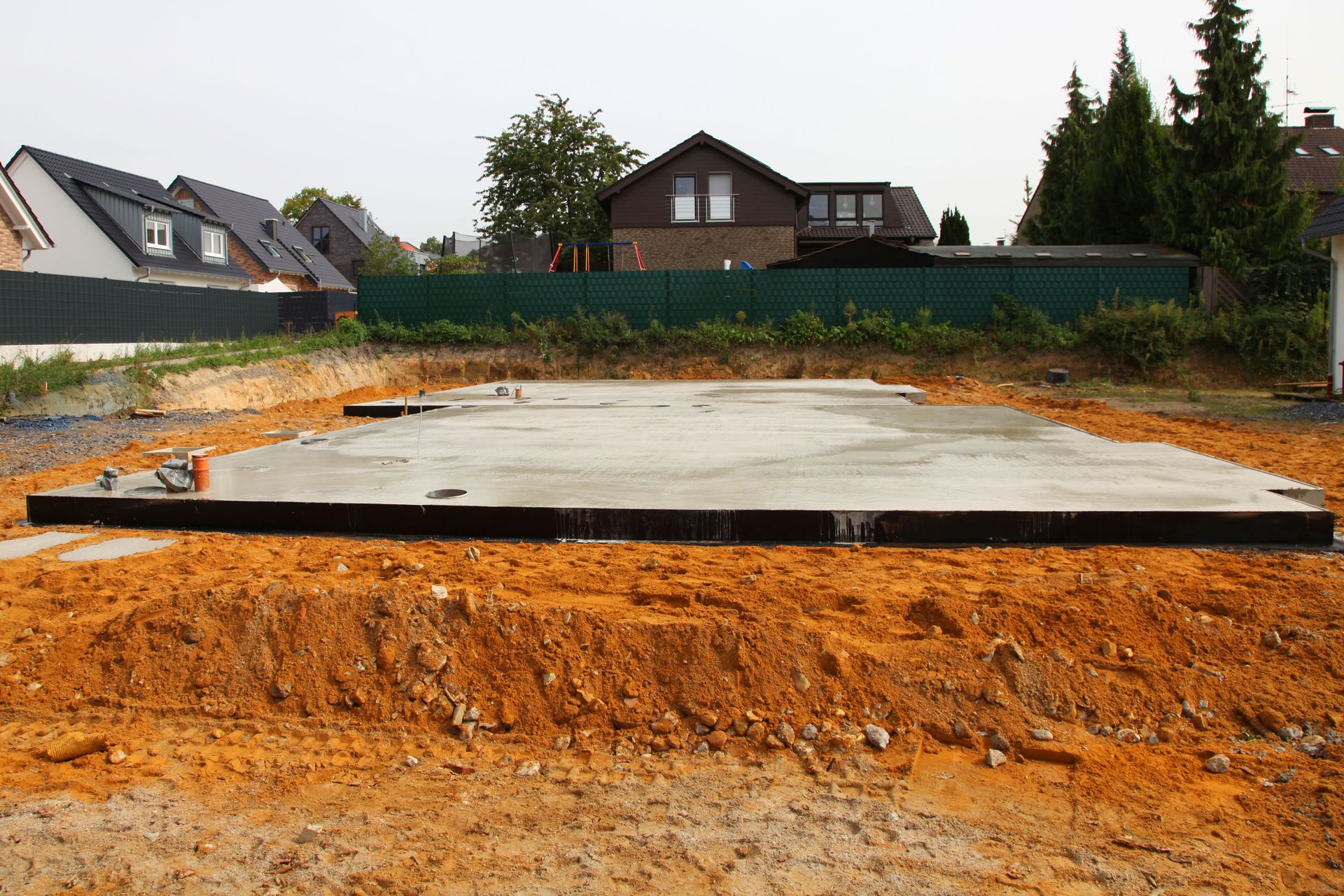 Newly poured concrete foundation for a building; surrounded by orange soil. Houses in the background.