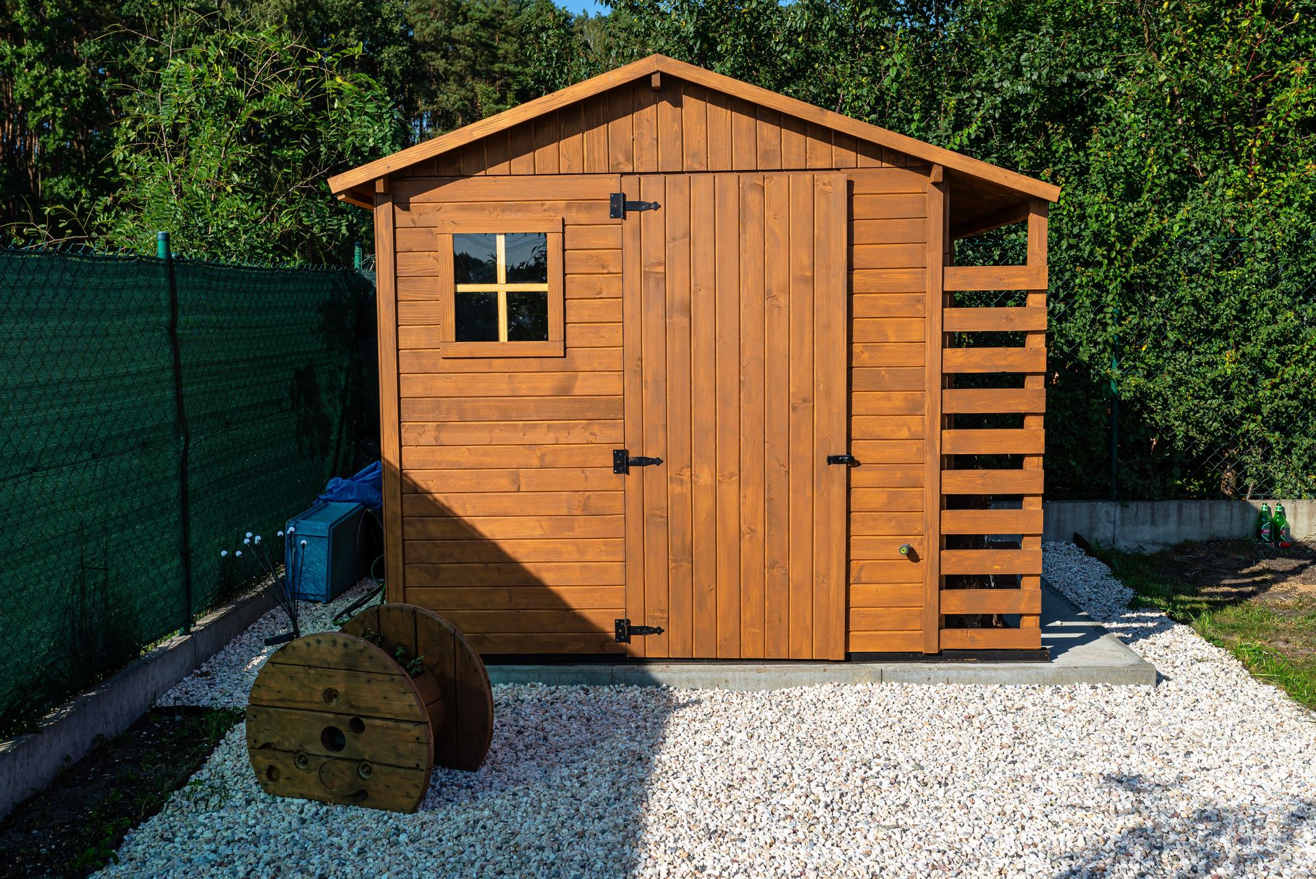 Wooden shed with doors, window, and ladder, on a gravel base, set against a green backdrop.