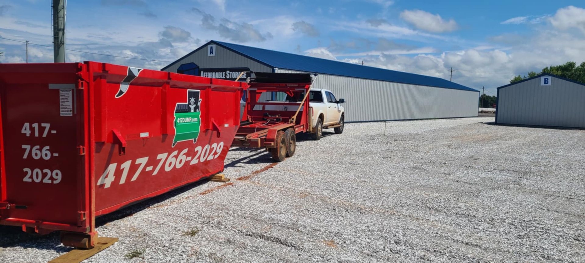 A red dumpster is sitting on top of a gravel lot next to a truck.