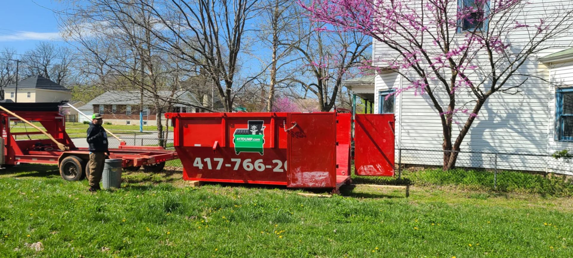 A red dumpster is parked in front of a white house.