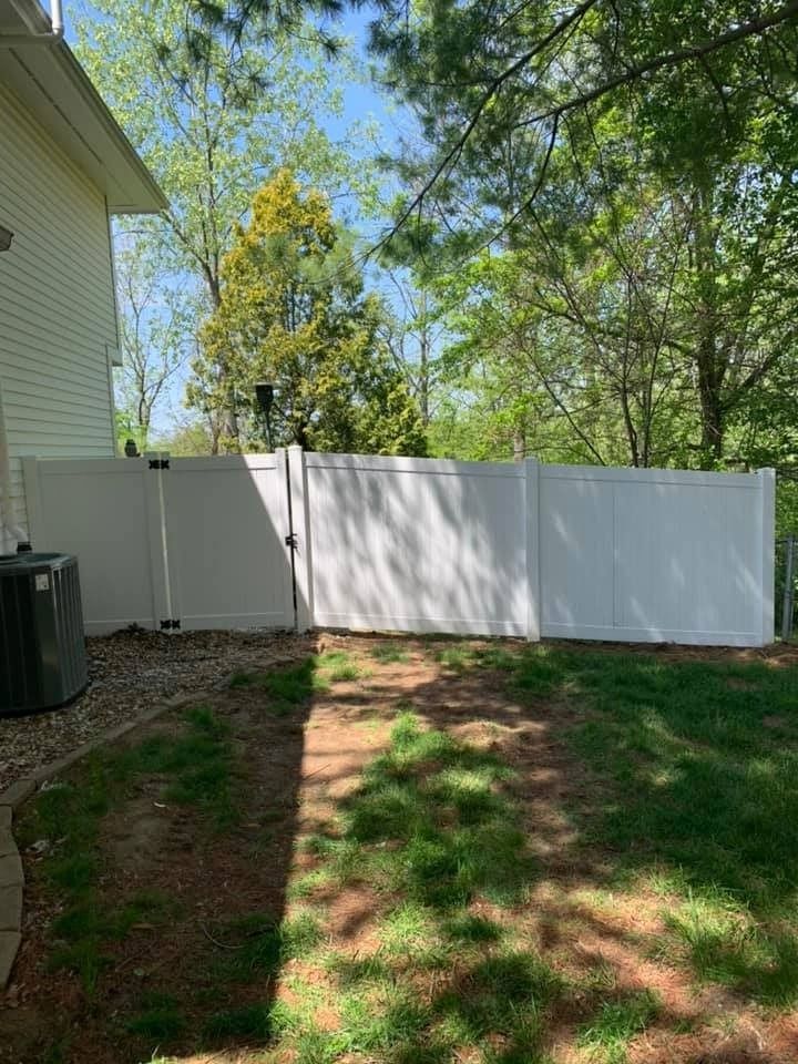 A white vinyl privacy fence sits in a backyard next to a house and air conditioning unit with trees in the background.