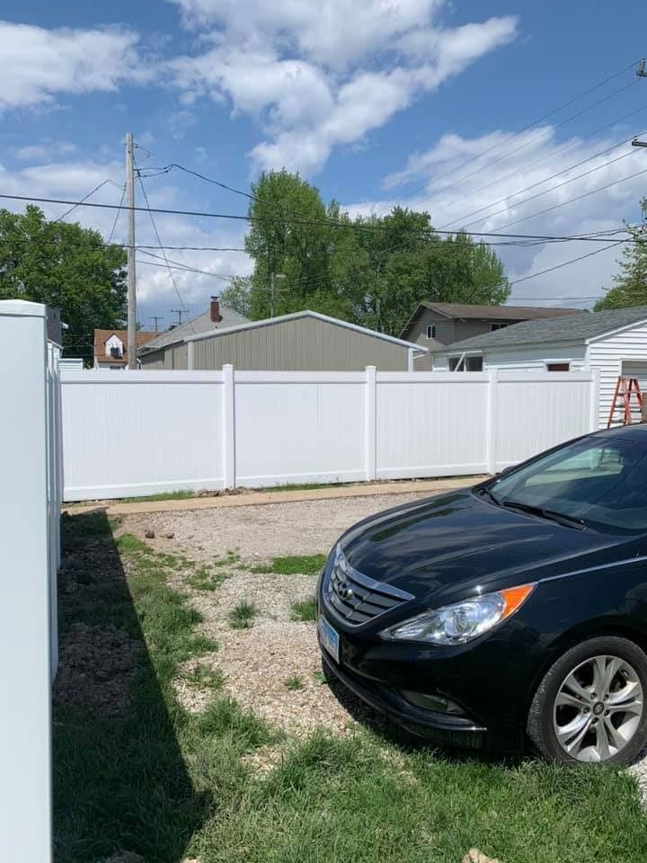 A black car parked on a gravel driveway in front of a white privacy fence with houses and trees in the background.