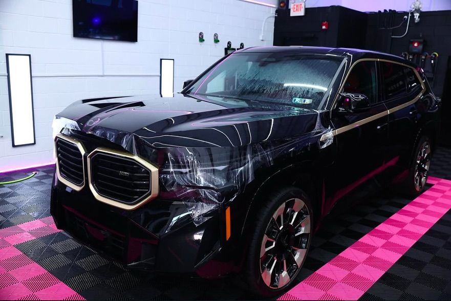 Black BMW XM SUV with gold trim parked in a detail shop with pink and black tiled flooring.
