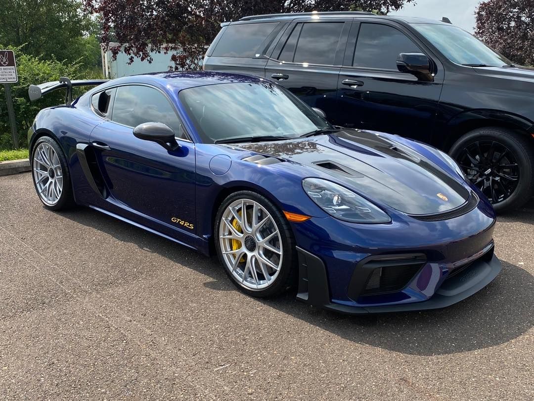 Blue Porsche 718 Cayman GT4 RS parked on pavement next to a black SUV, with a green tree in the background.