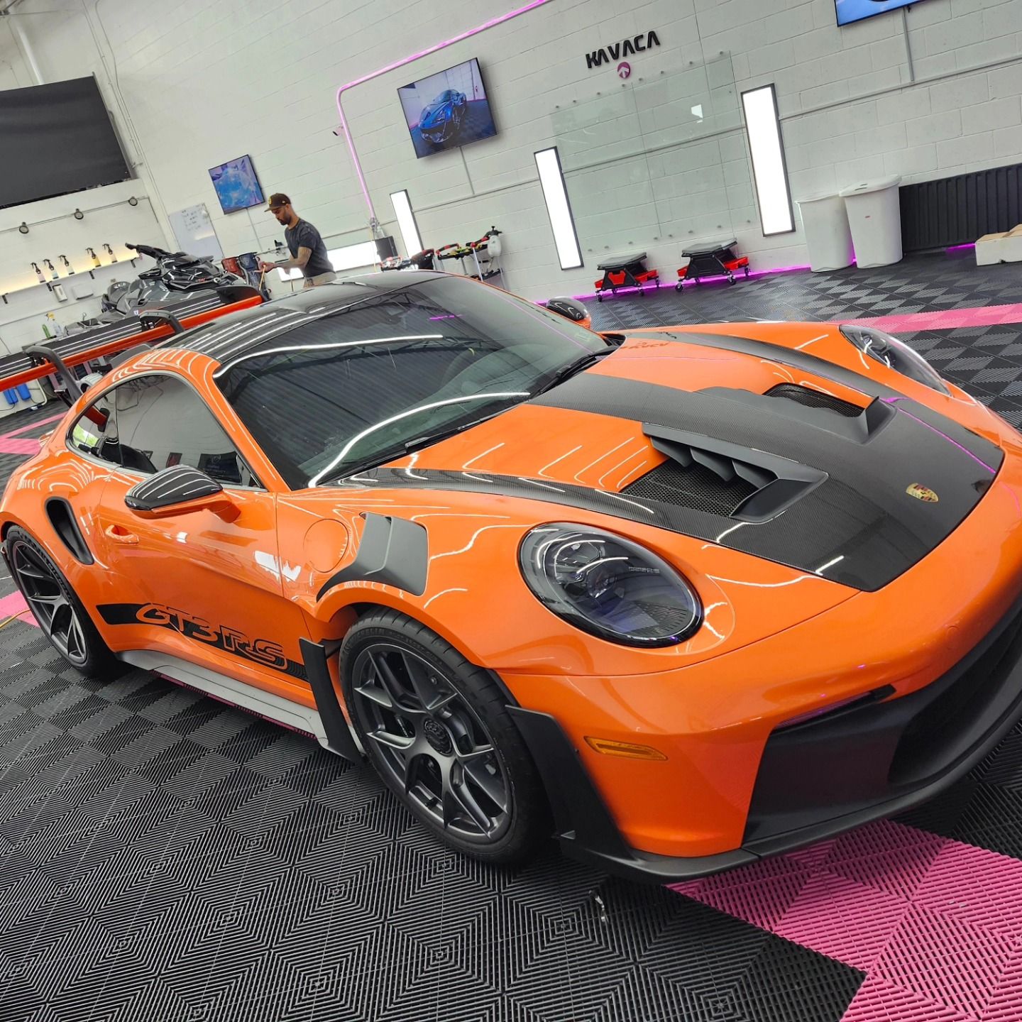 Orange Porsche 911 GT3 RS sports car with black accents in a detailing shop; a man works in the background.