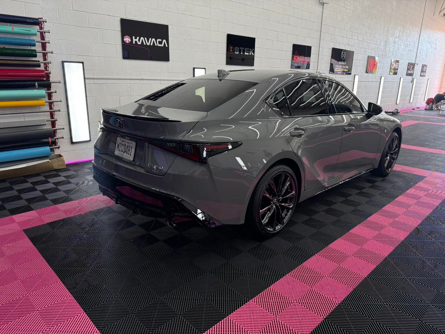 Gray Lexus sedan on a checkered black and pink floor, inside a shop, with rolls of colored vinyl.