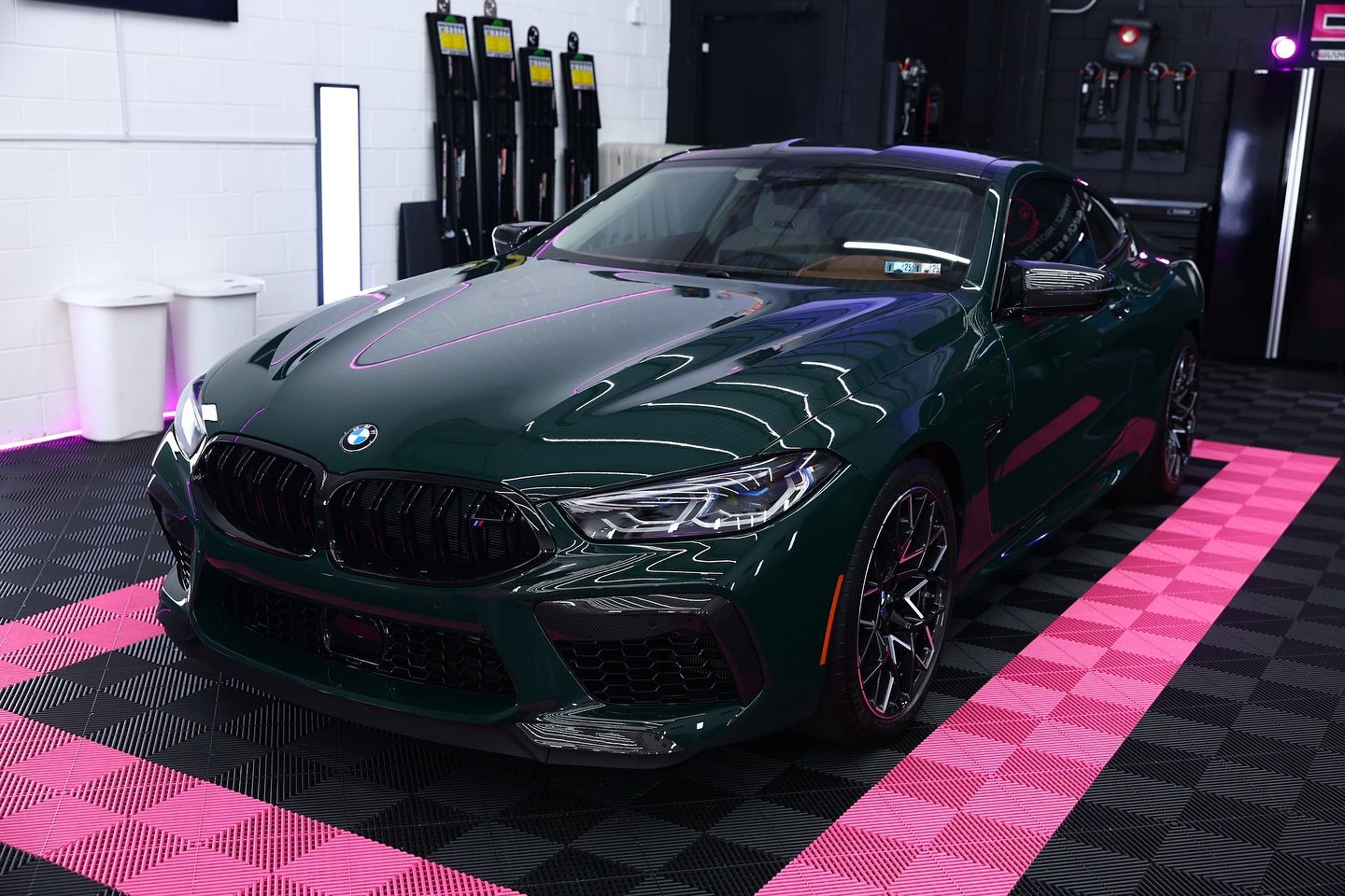 Dark green BMW M8 coupe parked inside a brightly lit garage with a pink and black checkered floor.