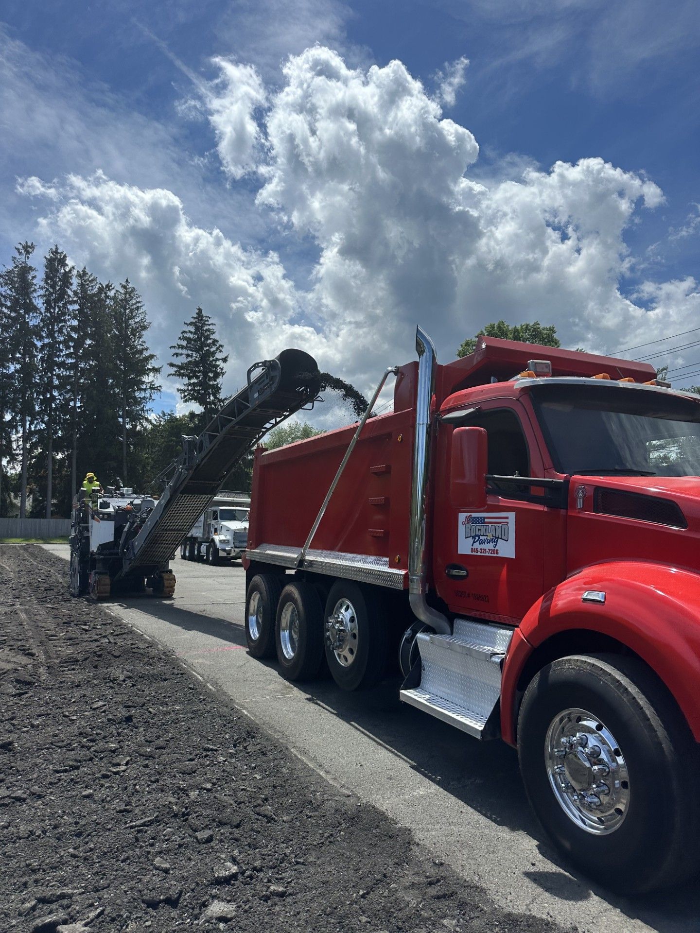 Road milling machine loading asphalt into a red dump truck on a sunny day.