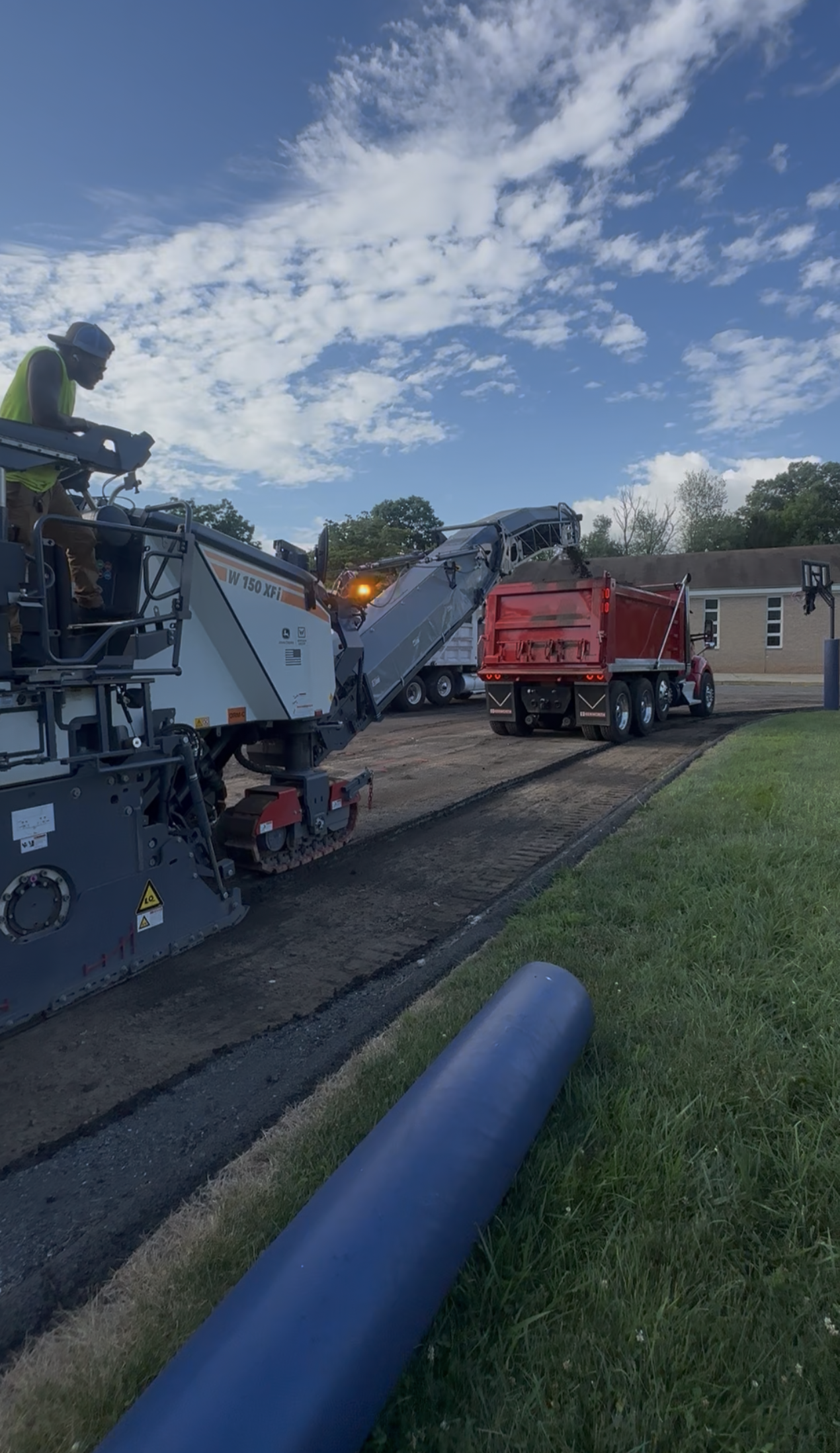 Asphalt paving in progress, a truck delivering asphalt to a paving machine on a road. A worker operating the machine.