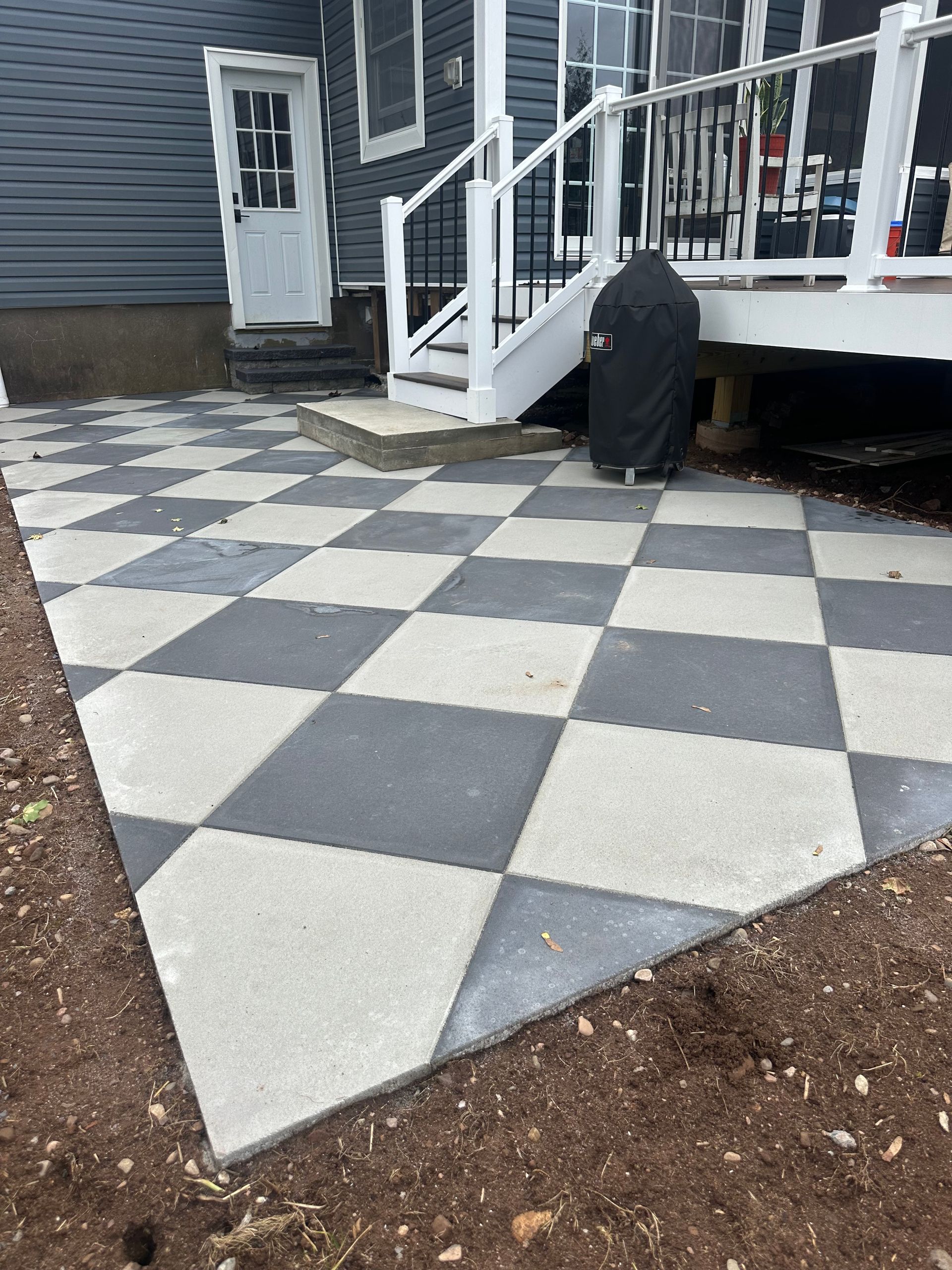 Checkerboard patio of gray and light pavers next to a house with a white door and railing.
