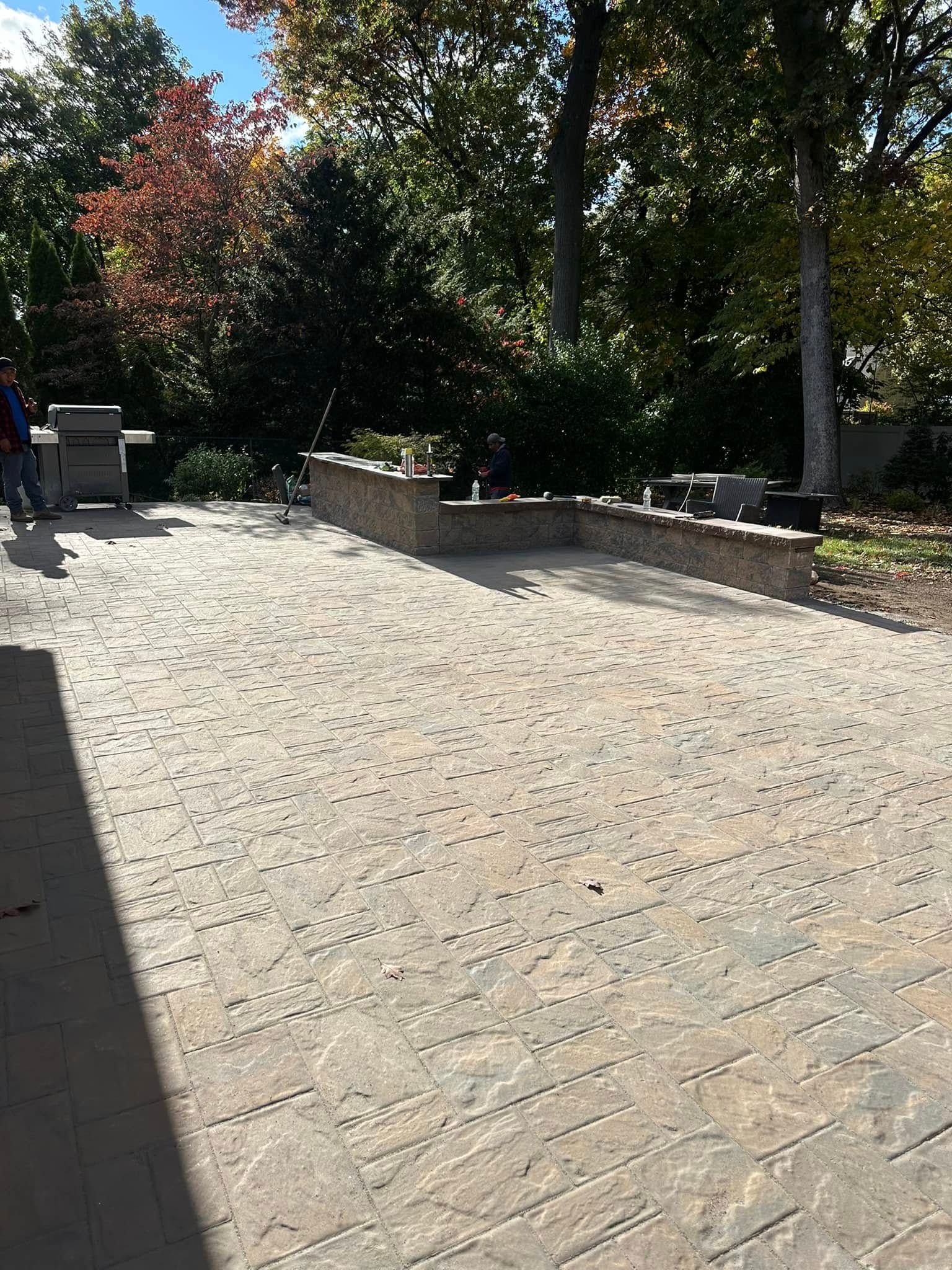 Outdoor patio with brick pavers, low brick wall, grill, and trees.