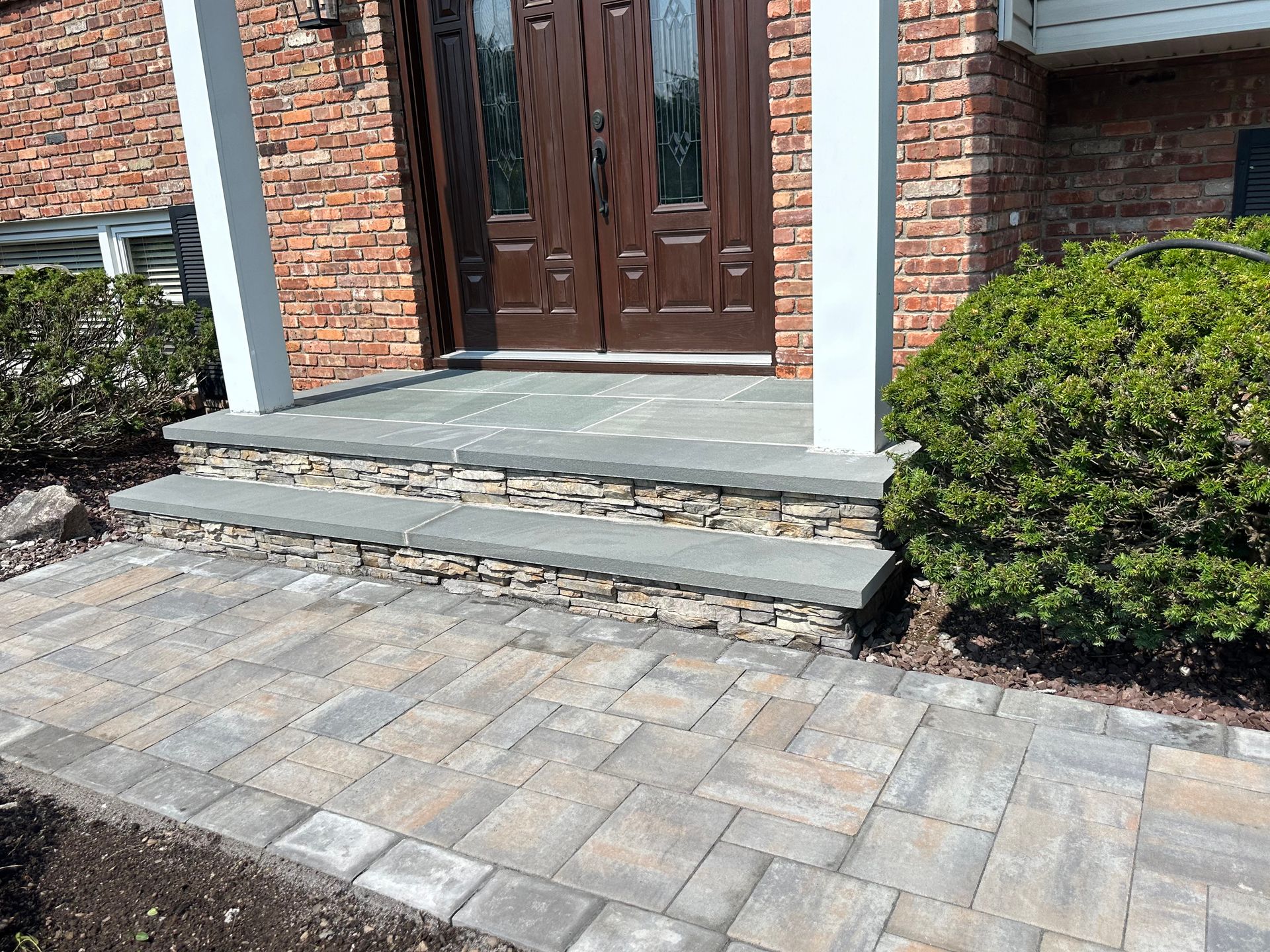 Stone walkway and steps leading to a brown door, with a green bush on the right and a brick facade.