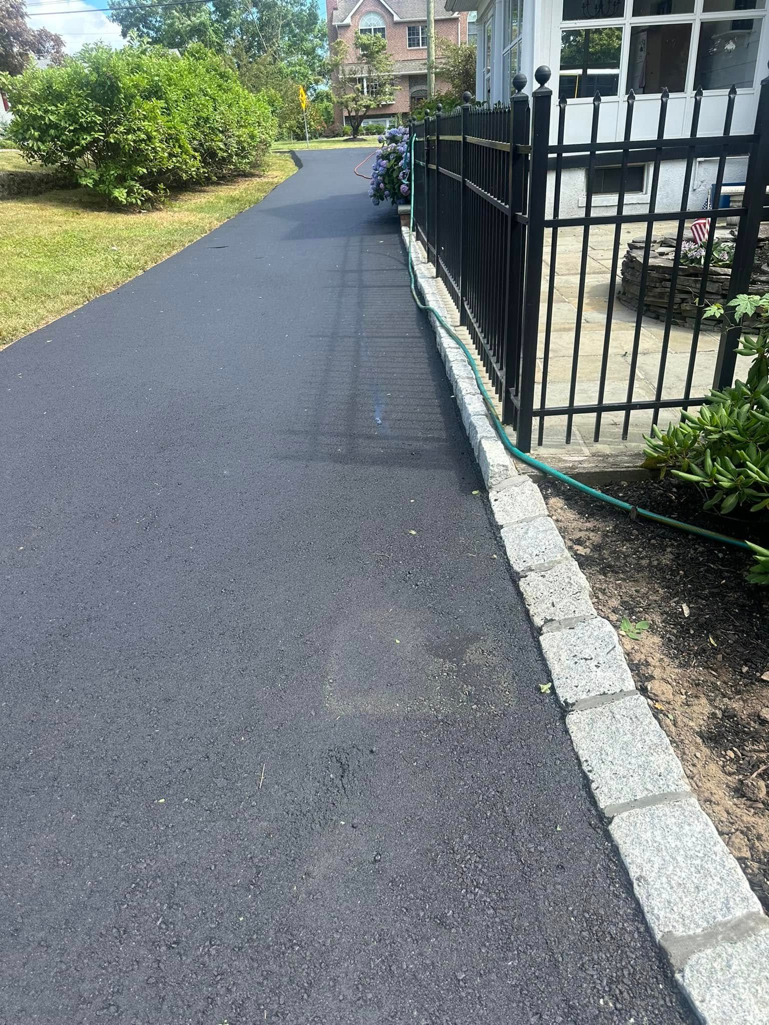 Asphalt driveway with stone edging and black metal fence. A garden and building are in the background.
