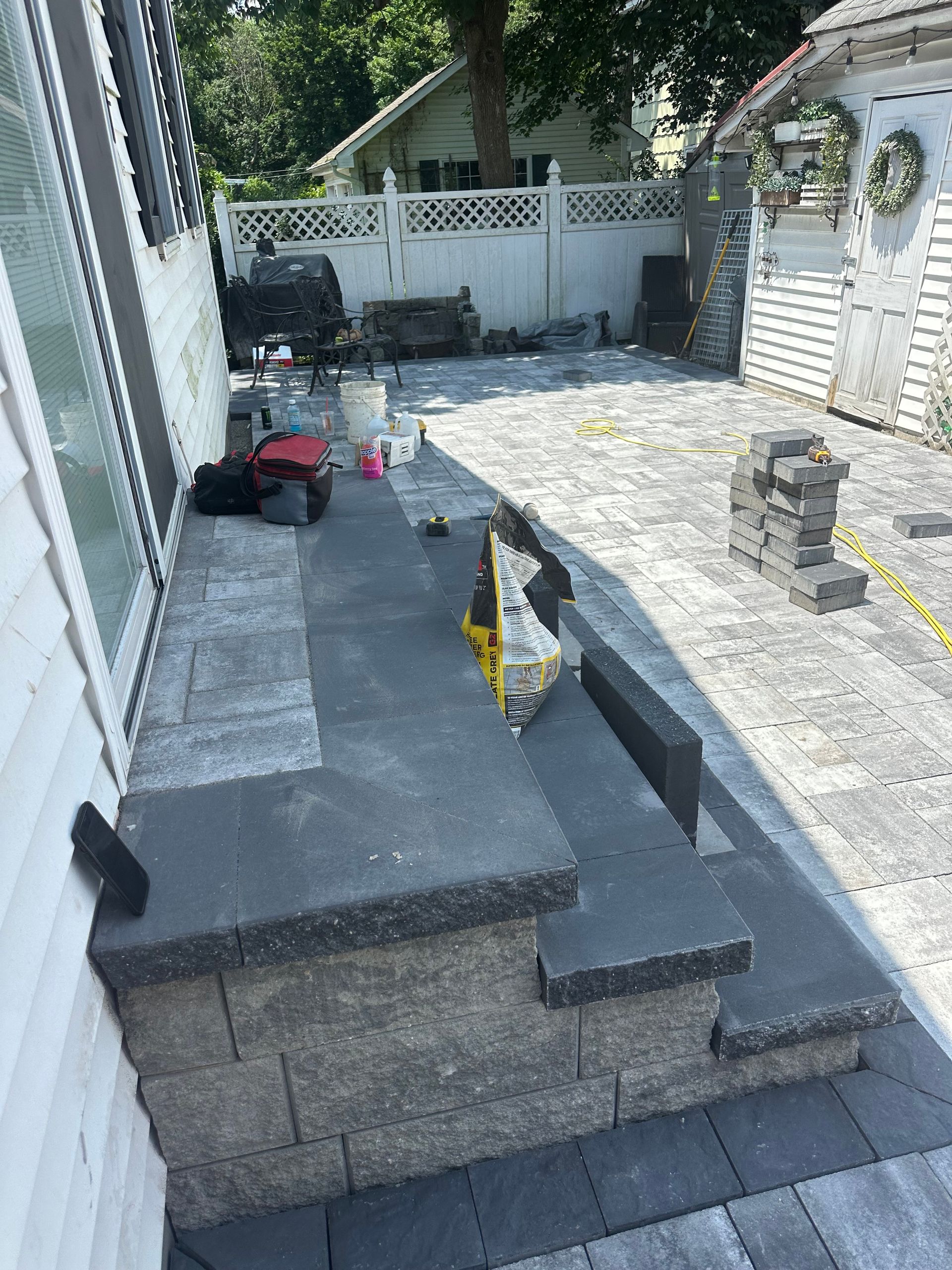 Stone patio and steps under construction next to a white house. Grey pavers and retaining wall blocks.
