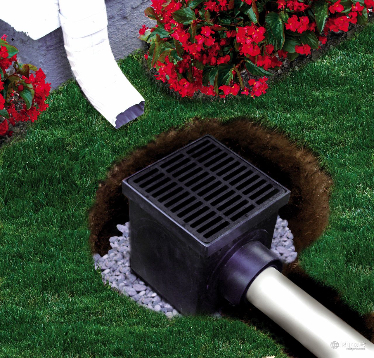 A black drainage grate installed in a grassy lawn, connected to a white downspout and an underground pipe.